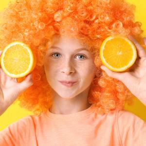 Image of a child wearing a bright orange afro wig with an orange in each hand.